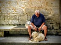 Aelterer Mann sitzt auf einer Steinbank vor einer Mauer mit einem Golden Retriever vor den Fuessen.jpg