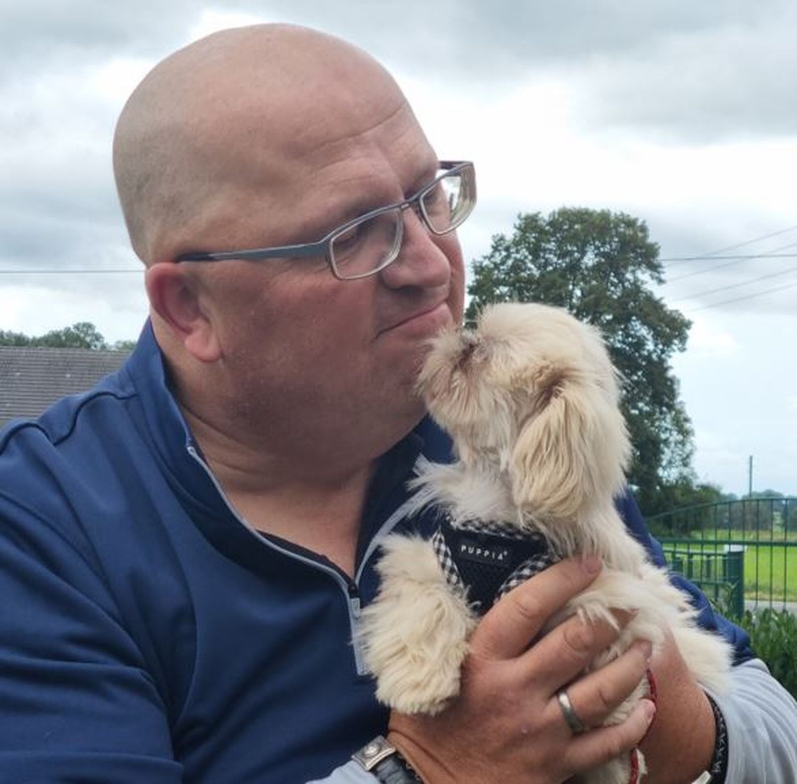 Ein Mann mit Glatze und Brille hält einen kleinen beigefarbenen Hund in Händen