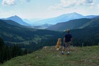 Ein Mann mit kurzer Hosen und blauen Shirt steht mit seinem Hund auf einem Berg und schaut aufs Bergpanorama.jpg