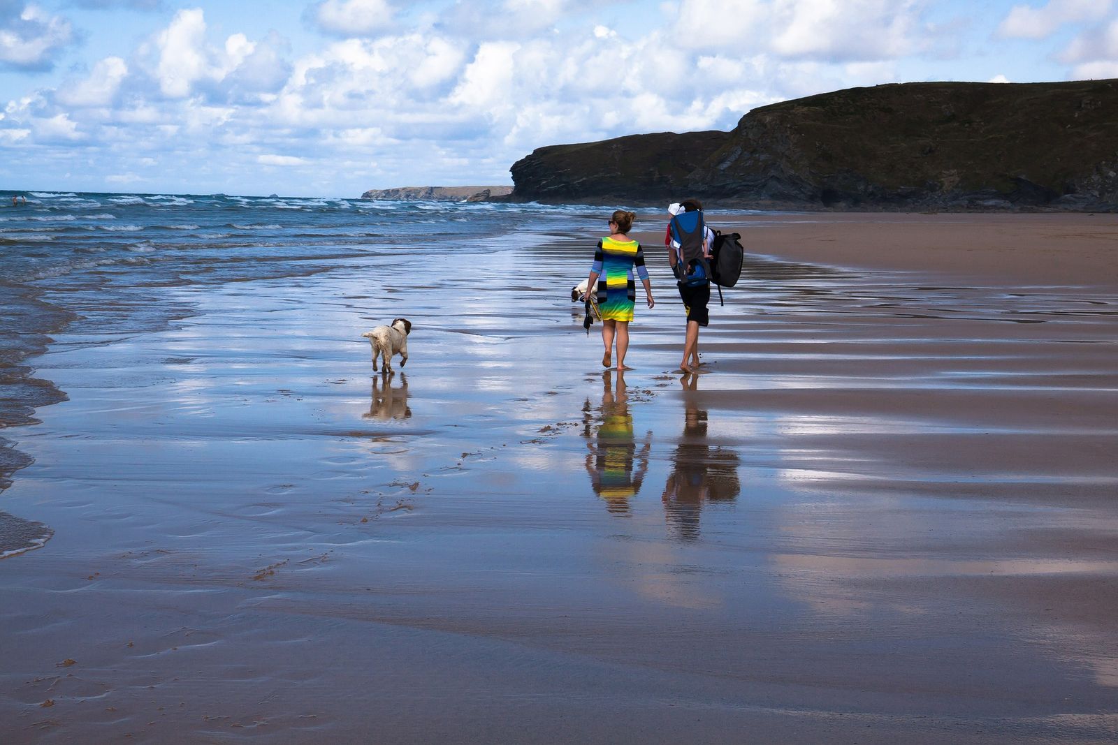 Ein Paar geht mit Kind im Rucksack und einem braun-weißen Hund am Strand spazieren