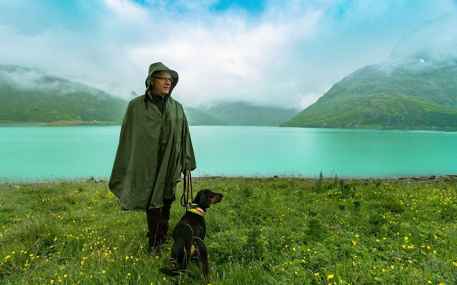 Ein Mann mit Brille und Regencape steht mit seinem angeleinten Jagdhund auf einer Wiese vor einem Bergsee und schaut nach rechts in die Ferne.